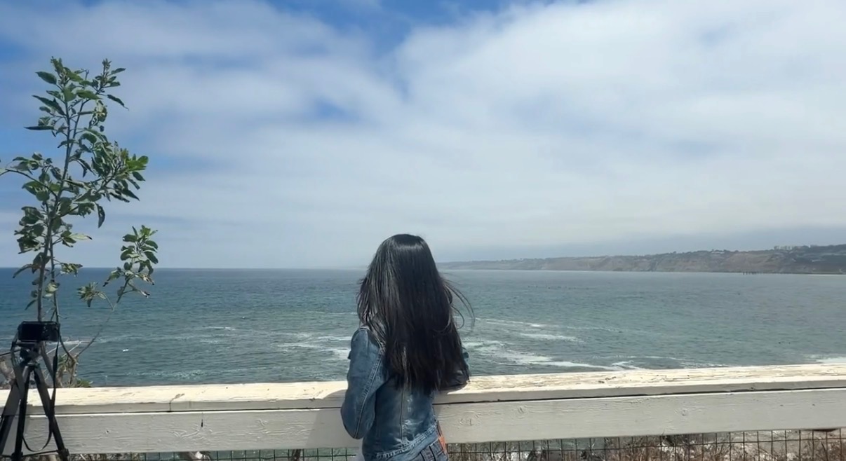 A person with long black hair in a denim jacket stands at a railing overlooking the ocean.