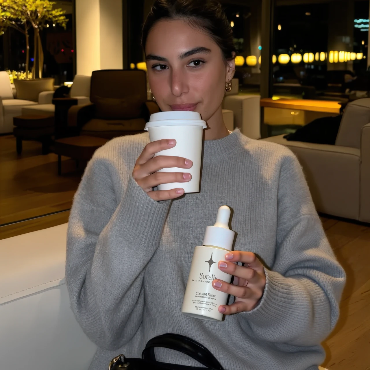 Person holding a coffee cup and a skincare product in a cozy room.