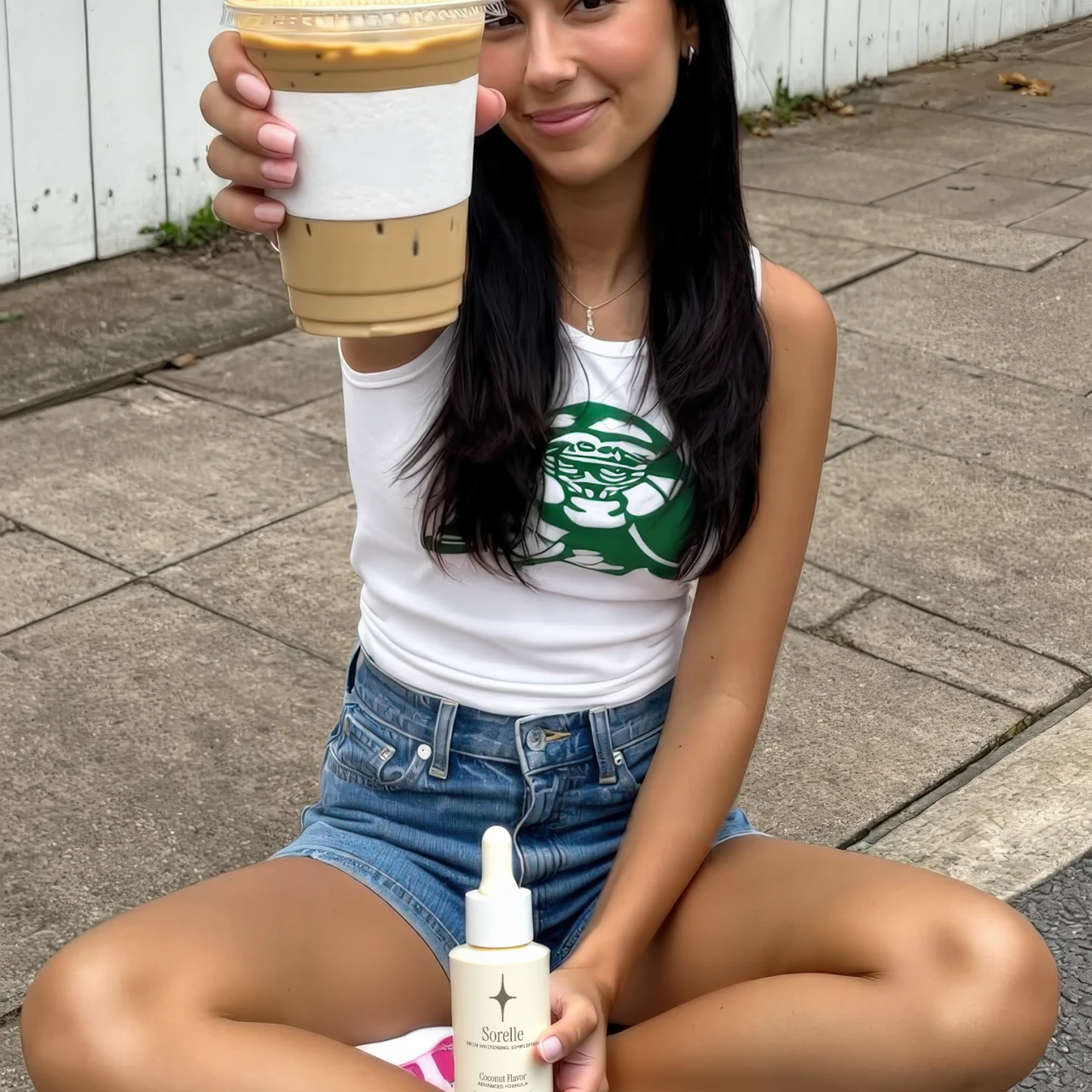 Woman sitting on pavement holding iced coffee and skincare product.