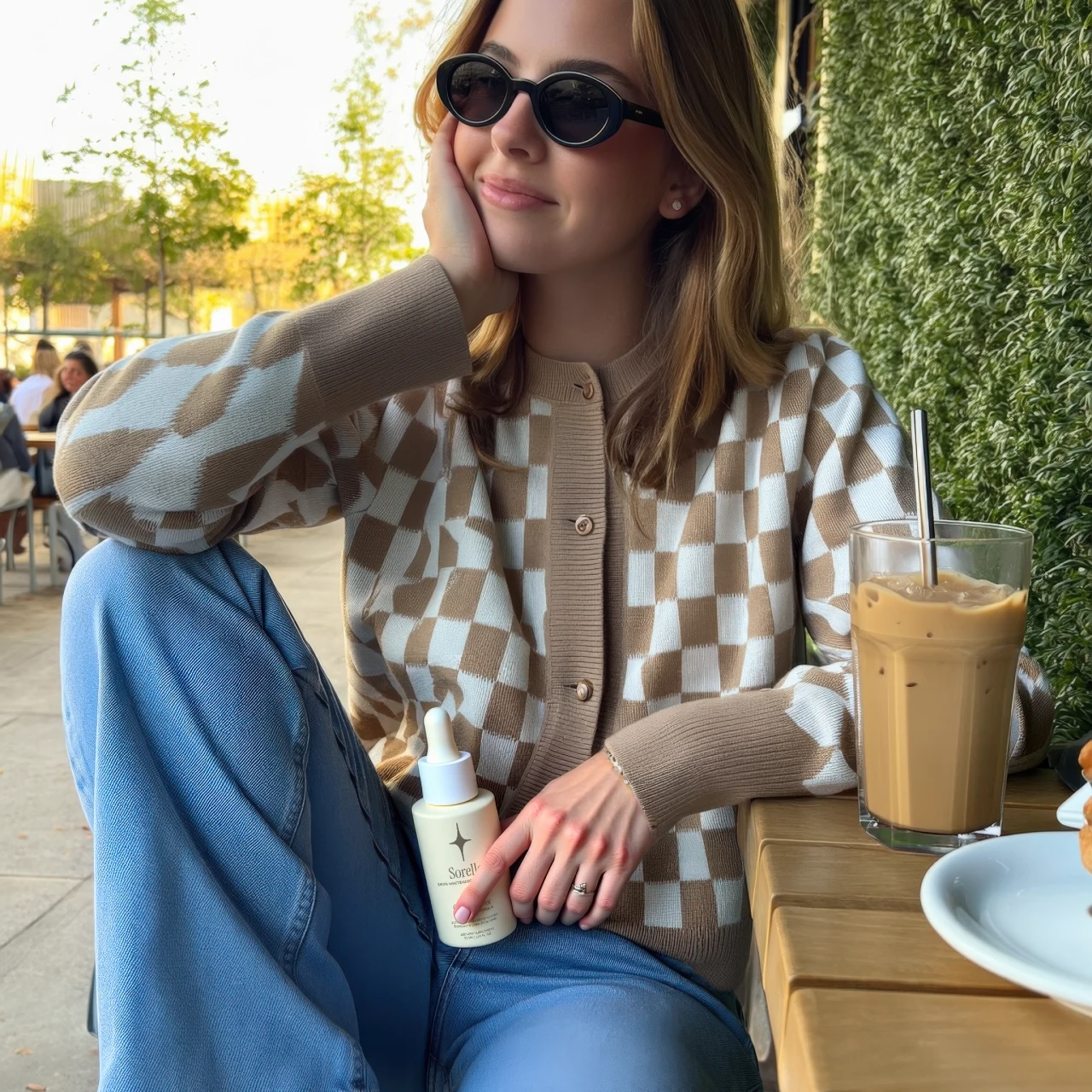 Woman in checkered sweater holding a bottle, sitting with iced coffee outdoors.