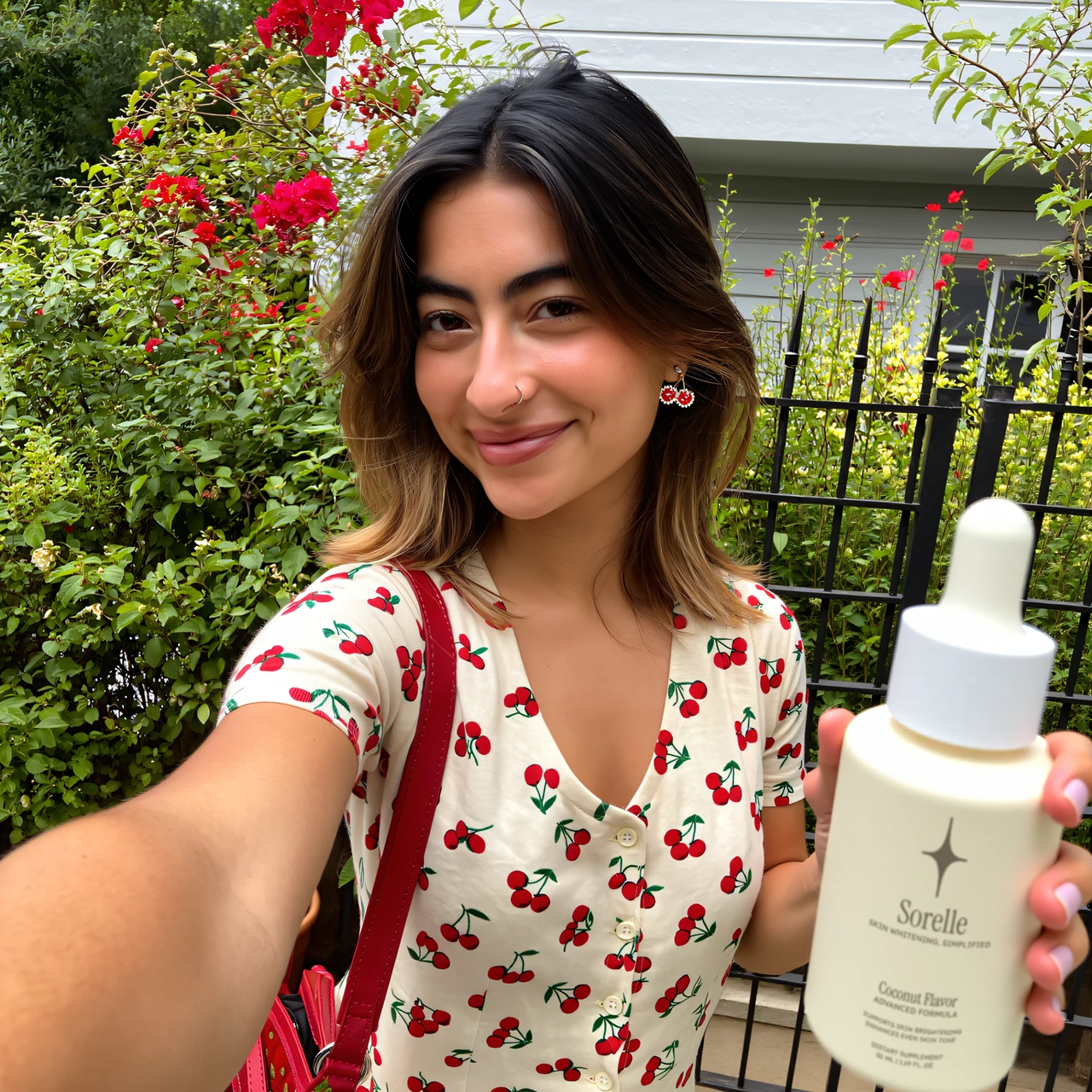 Person holding a bottle, wearing a cherry-patterned shirt, standing in front of a garden.