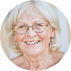 Elderly woman with glasses smiling, wearing a light top, against a neutral background.