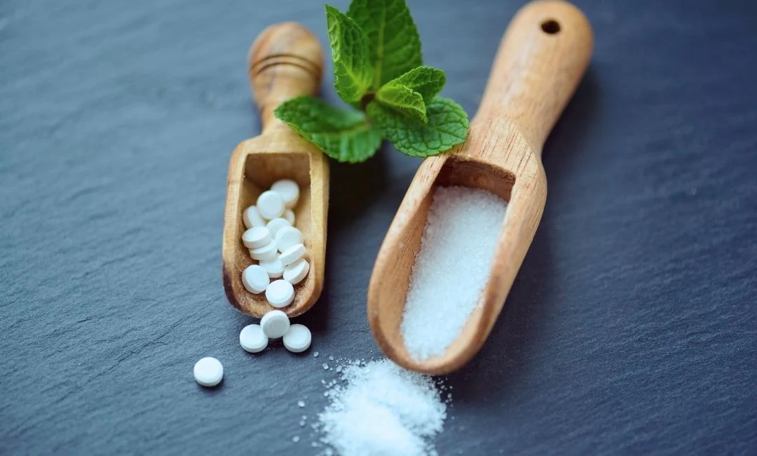 Wooden scoops with white tablets, granulated substance, and mint leaves on a dark surface.