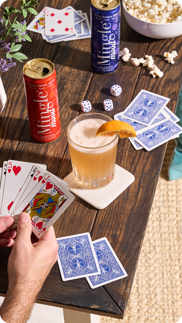 A person plays cards on a wooden table with Mingle Mood drinks, a cocktail, popcorn, and dice.