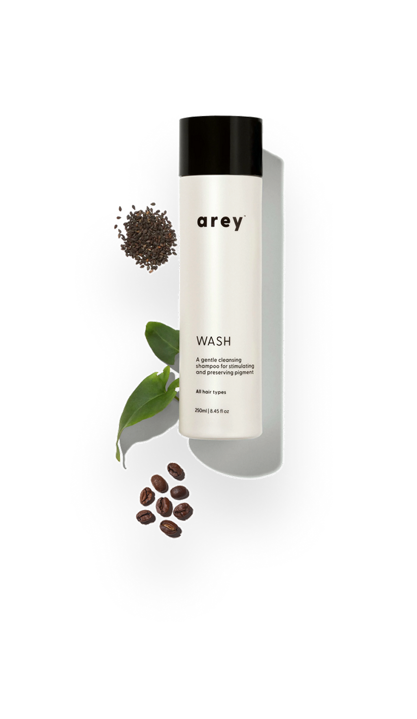 A bottle of Arey Wash shampoo on a white background with black sesame seeds, green leaves, and coffee beans.
