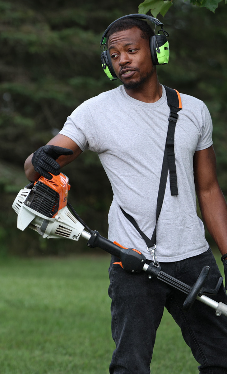 Person wearing ear protection, holding a gas-powered trimmer in a garden setting.