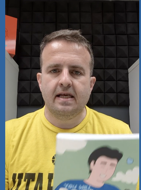 Man in a yellow shirt holding a book indoors.