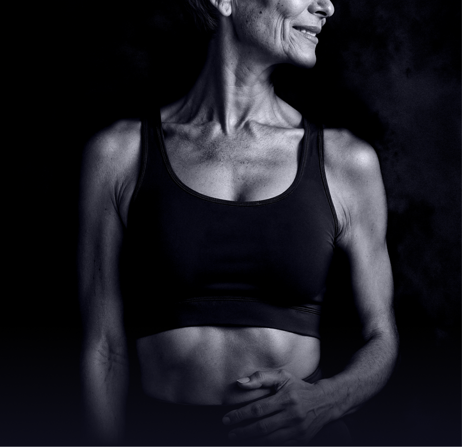 A black and white photograph of a fit, older woman in a sports bra against a dark background.