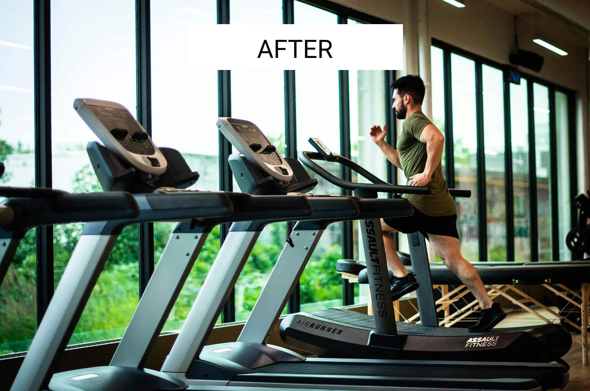 Man running on a treadmill in a gym with large windows.