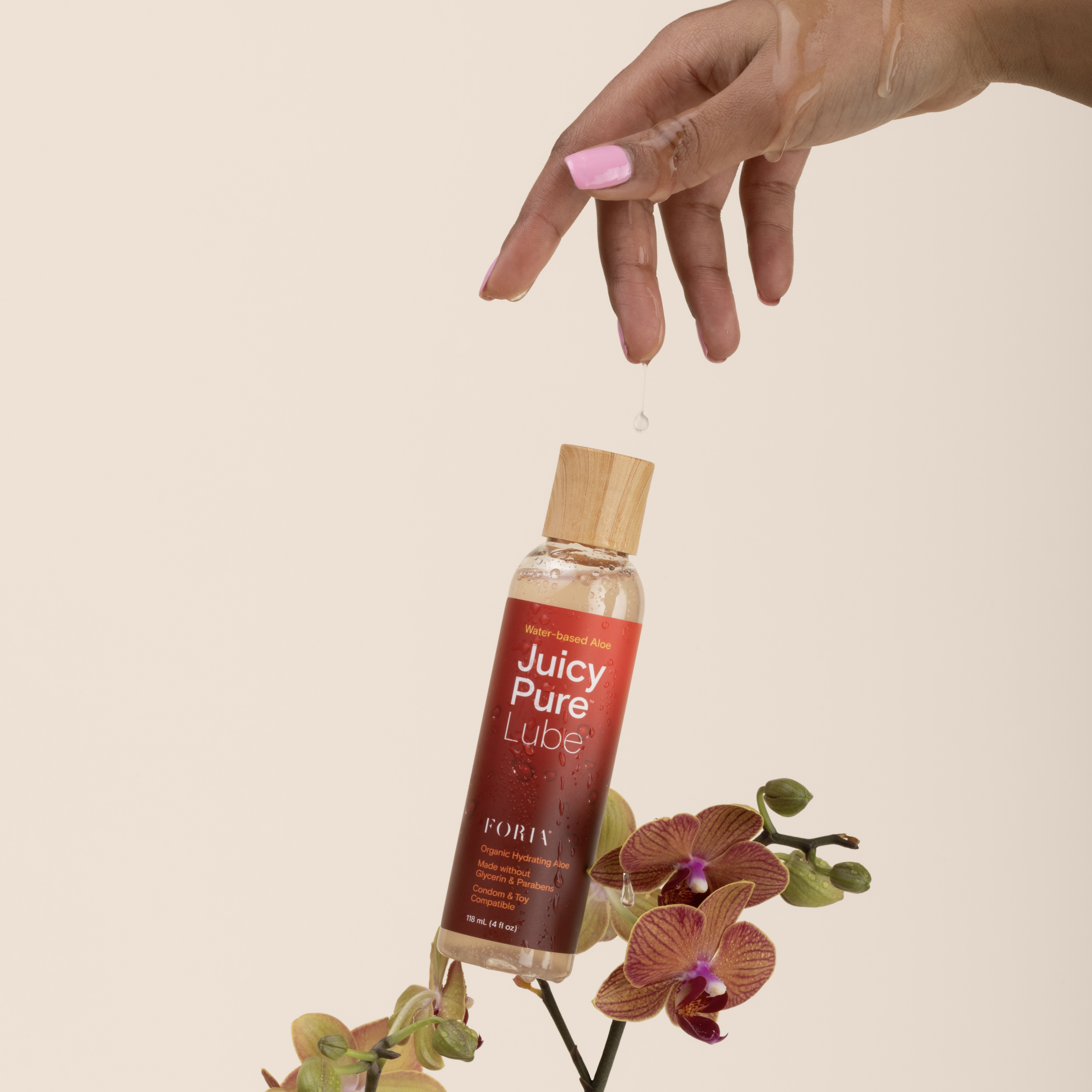 A hand with pink fingernails drips a clear liquid onto a bottle of lubricant resting on an orchid.