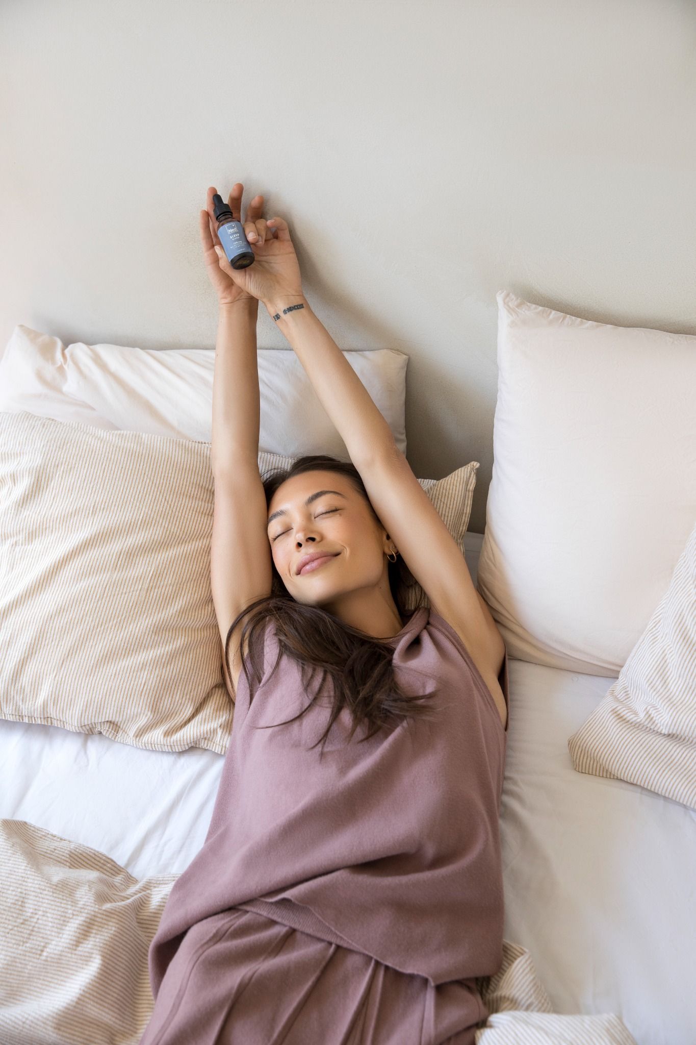 An overhead shot of a woman in bed, stretching with eyes closed while holding a small bottle.