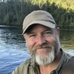 A man with a graying beard and a tan baseball cap smiling, with a lake and trees behind him.