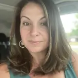 A woman with shoulder-length brown hair and highlights looks at the camera from inside a car.