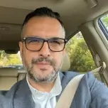 A man with glasses and a beard, wearing a suit jacket, takes a selfie inside a car.