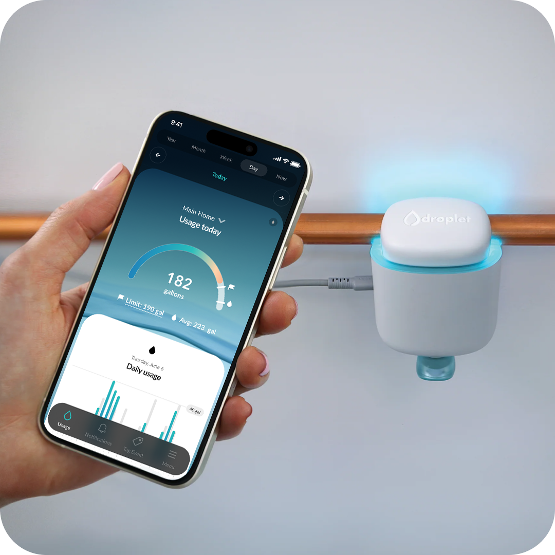 Hand holding a smartphone showing water usage app, near a water sensor on a pipe.