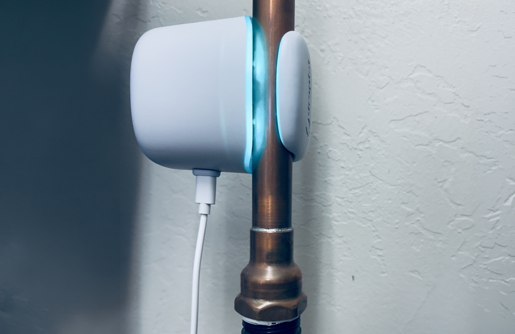 A white electronic device with a glowing blue light is clamped onto a copper pipe against a white wall.