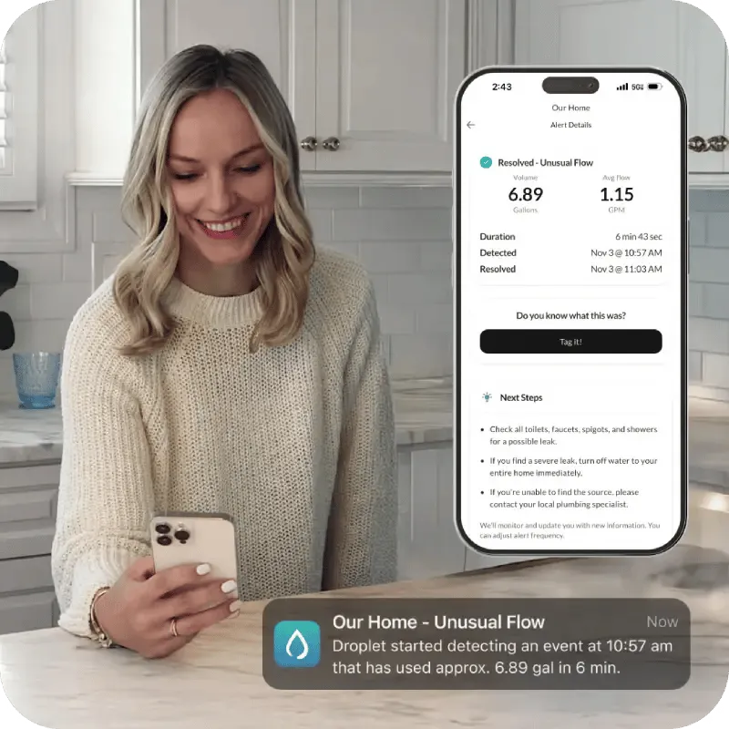 A woman smiles while looking at a smart home water usage alert on her phone in her kitchen.
