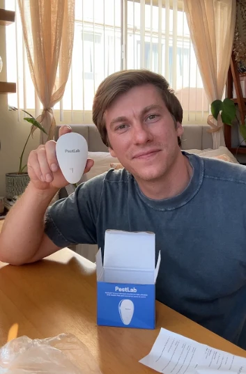 A smiling man sits at a table holding a white PestLab device, with the product box in front.