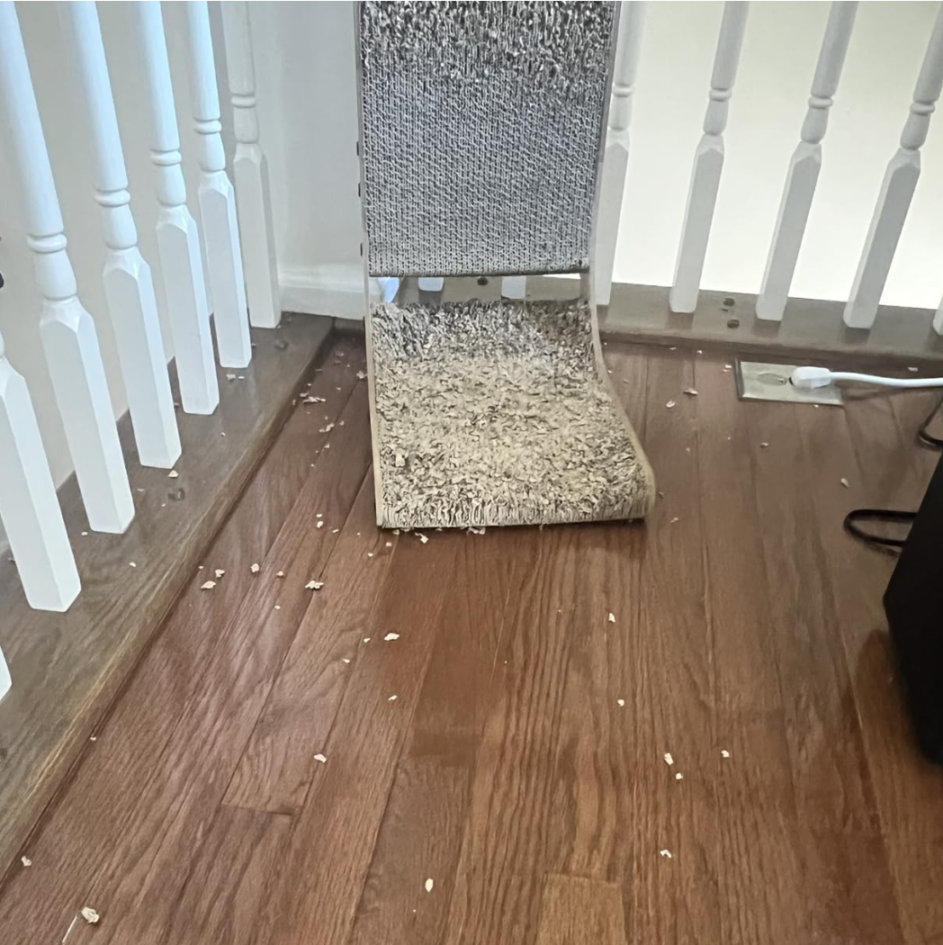 Worn-out cat scratching post with debris on wooden floor near white railing.