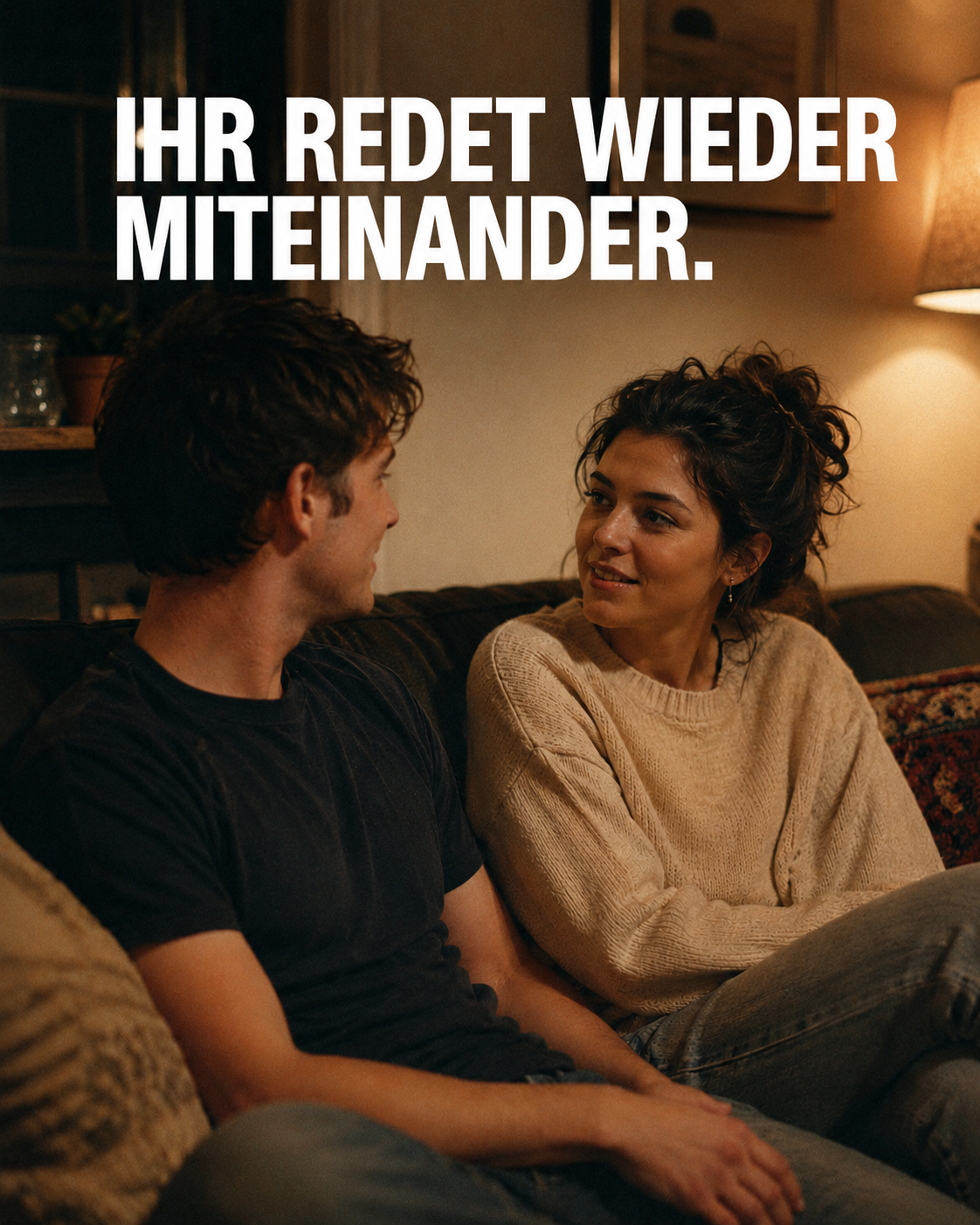 A young man and woman sit on a couch in a warmly lit room, talking to each other.