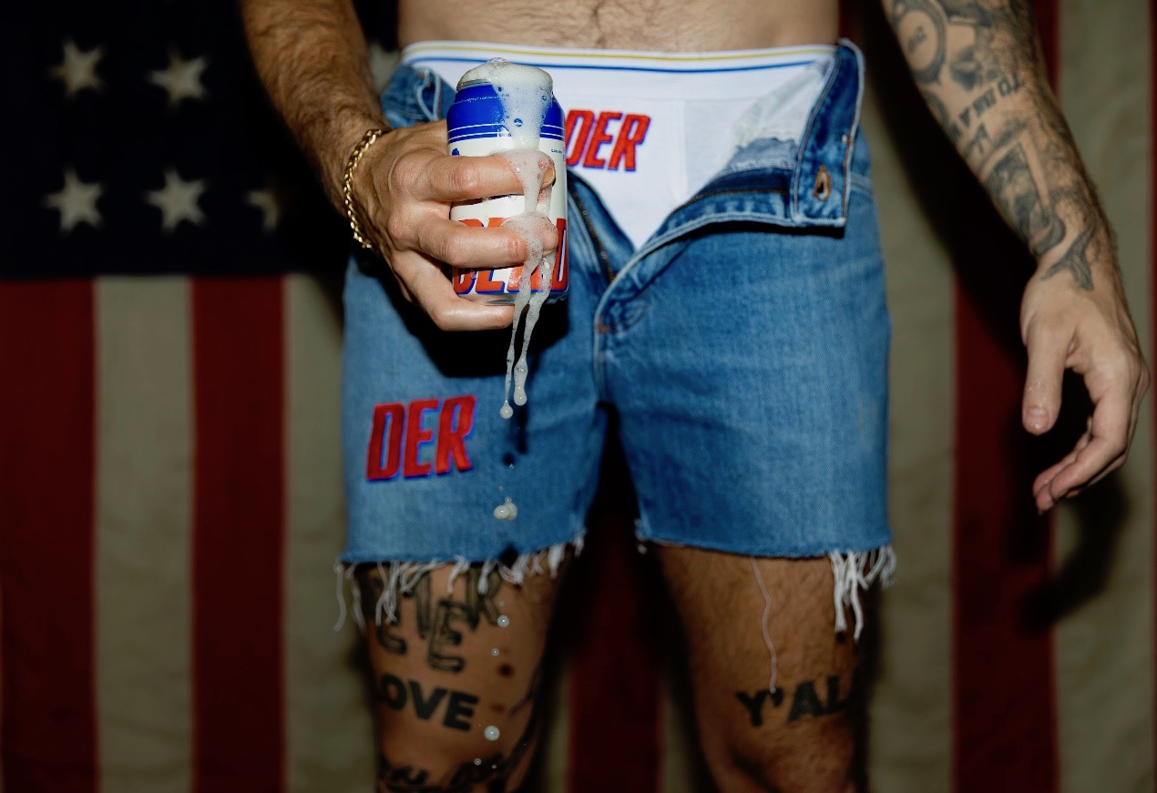Man in denim shorts holding a frothy can, tattoos on legs.