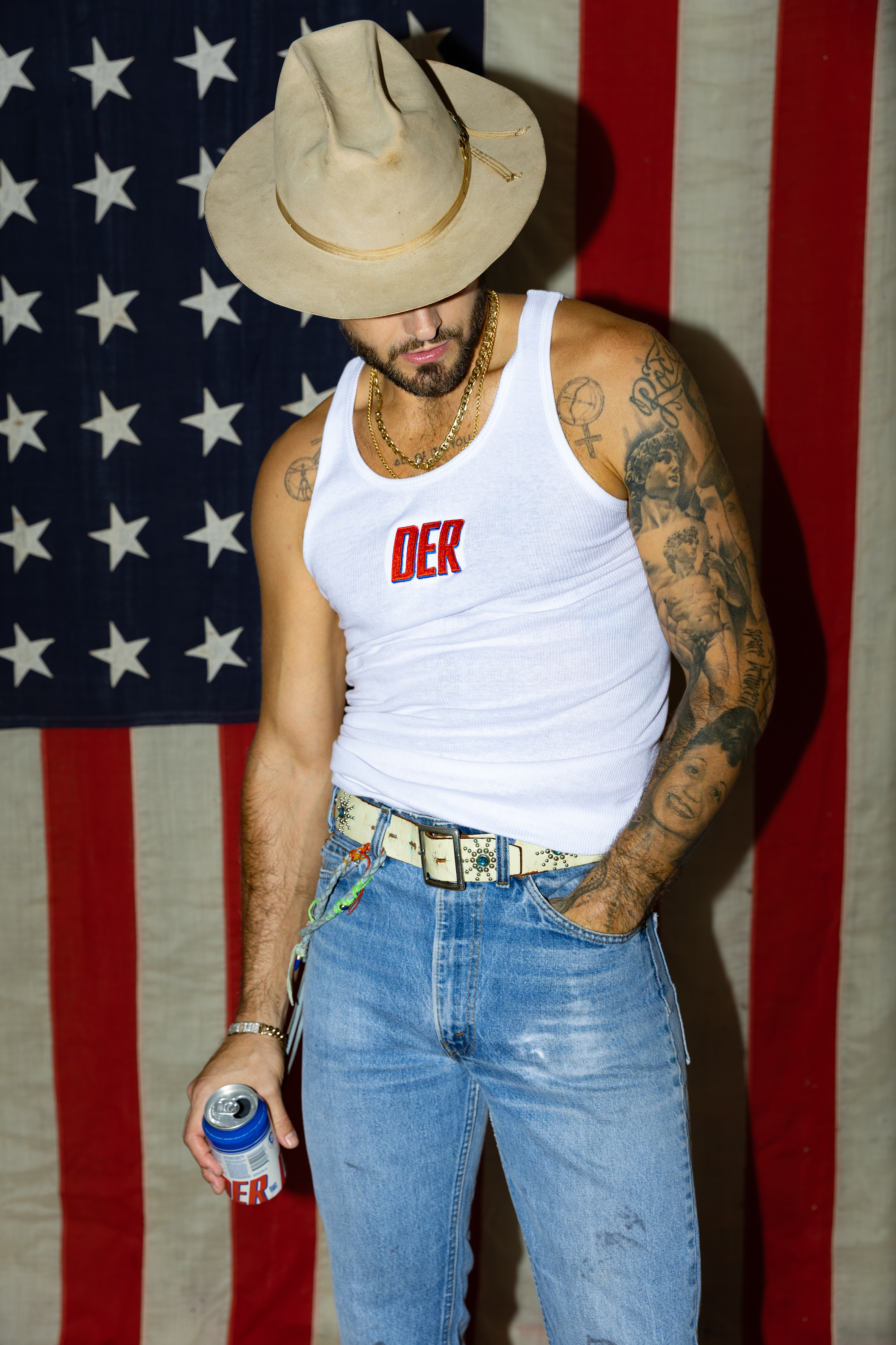 Person in hat and tank top, holding a can, standing before an American flag.