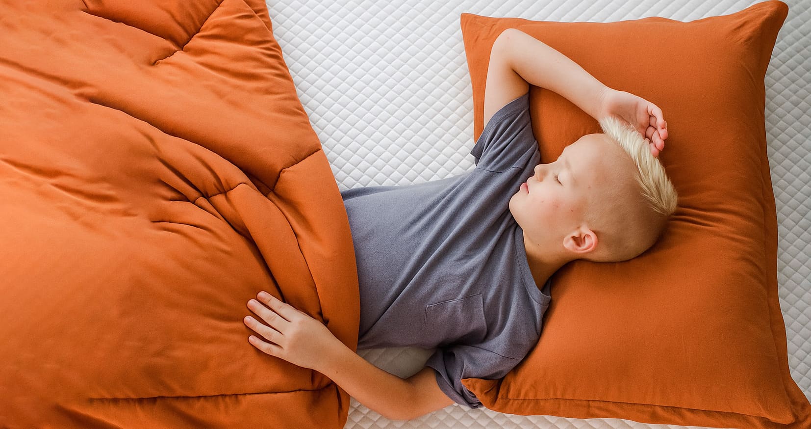 Child sleeping on a bed with an orange pillow and comforter, lying on a white quilted mattress.