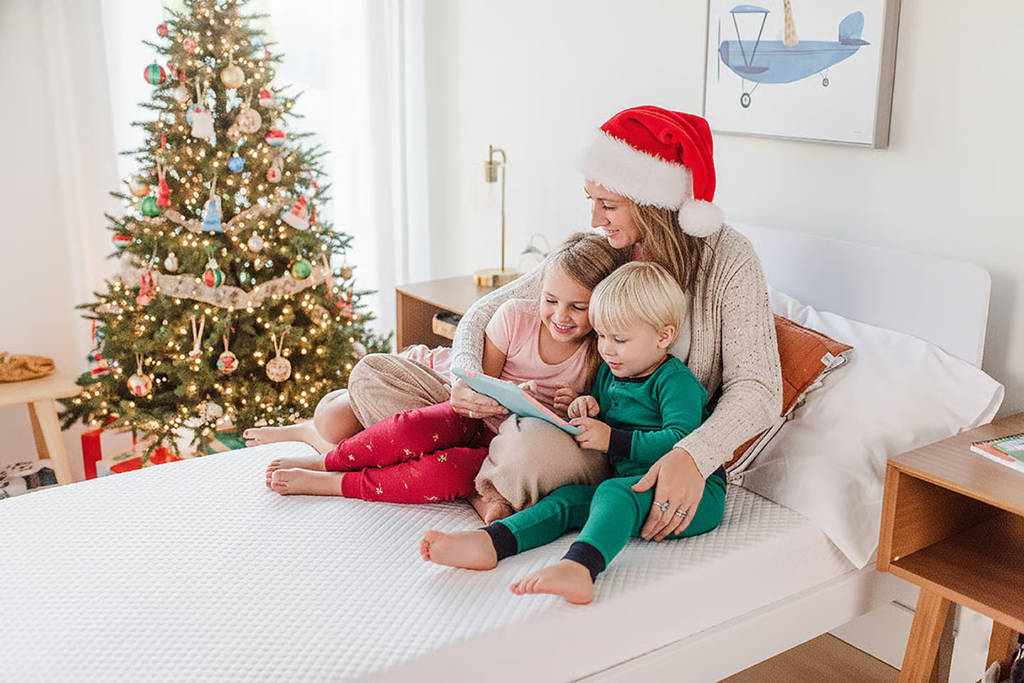 Woman wearing Santa hat and reading to her 2 school-aged children on top of white mattress.