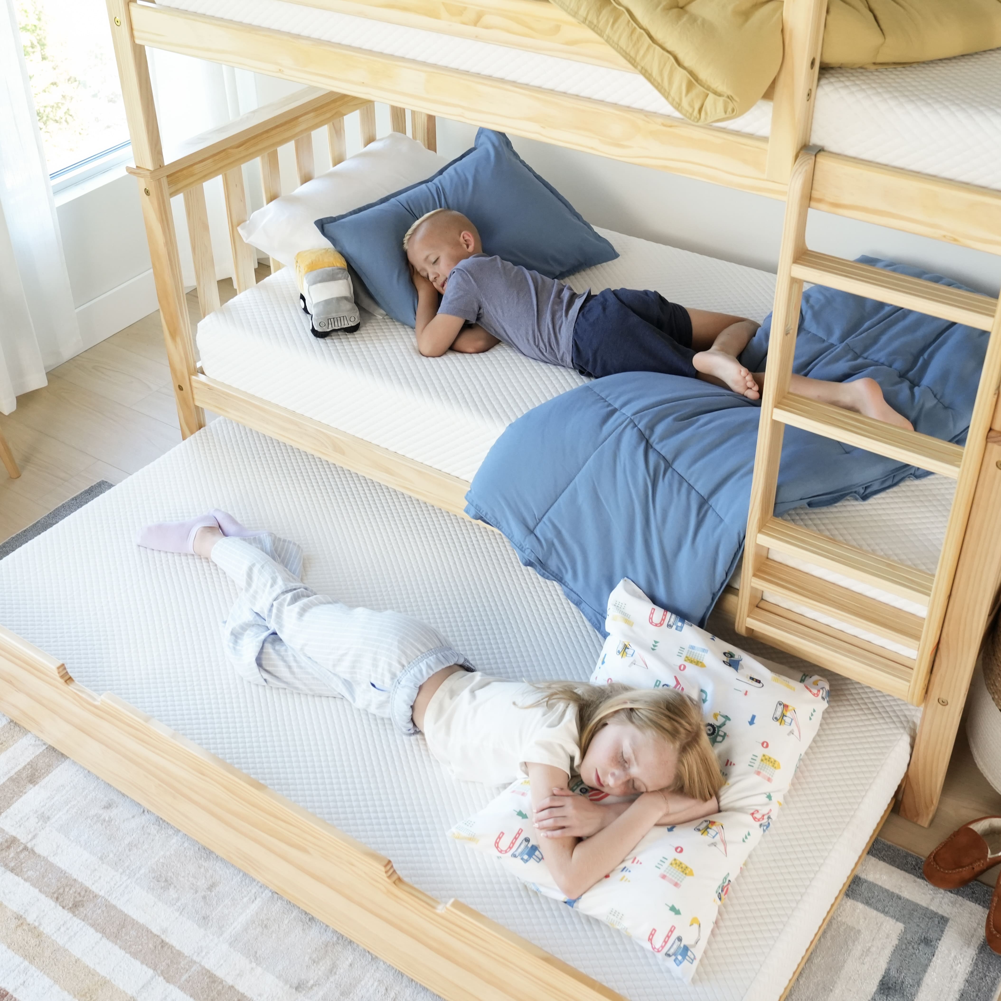 Two children sleeping on wooden bunk beds, one on the middle bunk with blue bedding and one on the pull-out trundle with a patterned pillow.