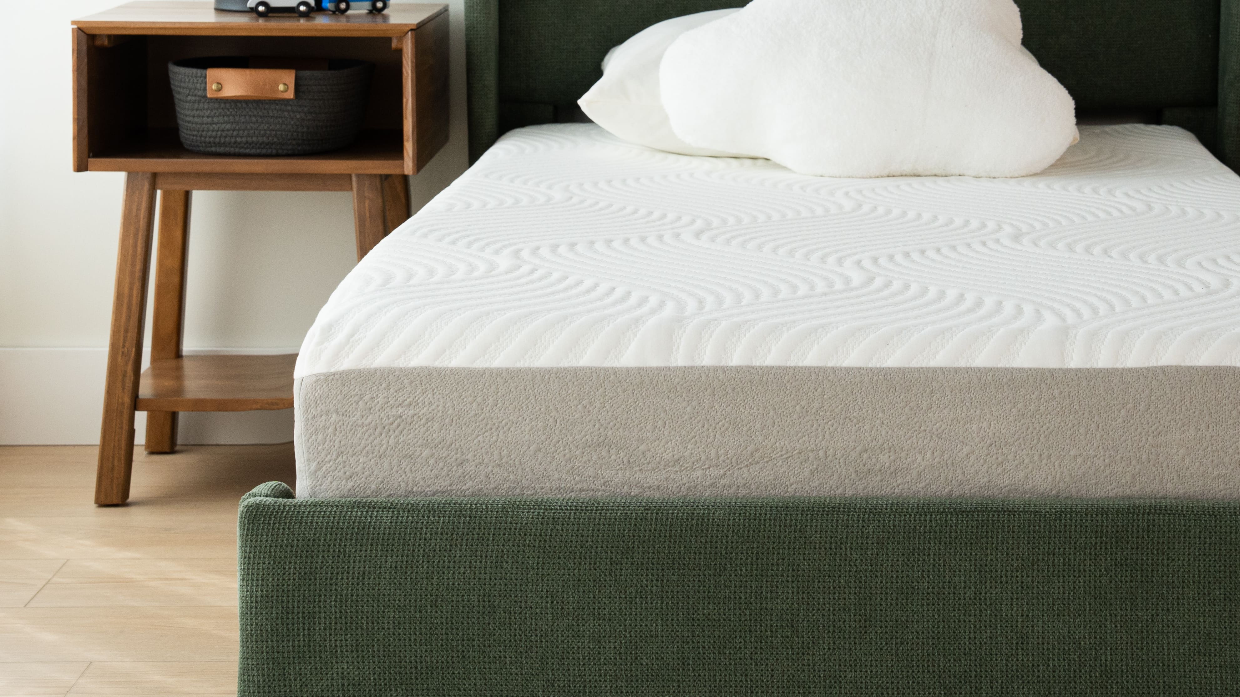 A white and grey mattress on a green upholstered bed with a white pillow. A bed-side table is next to the bed.