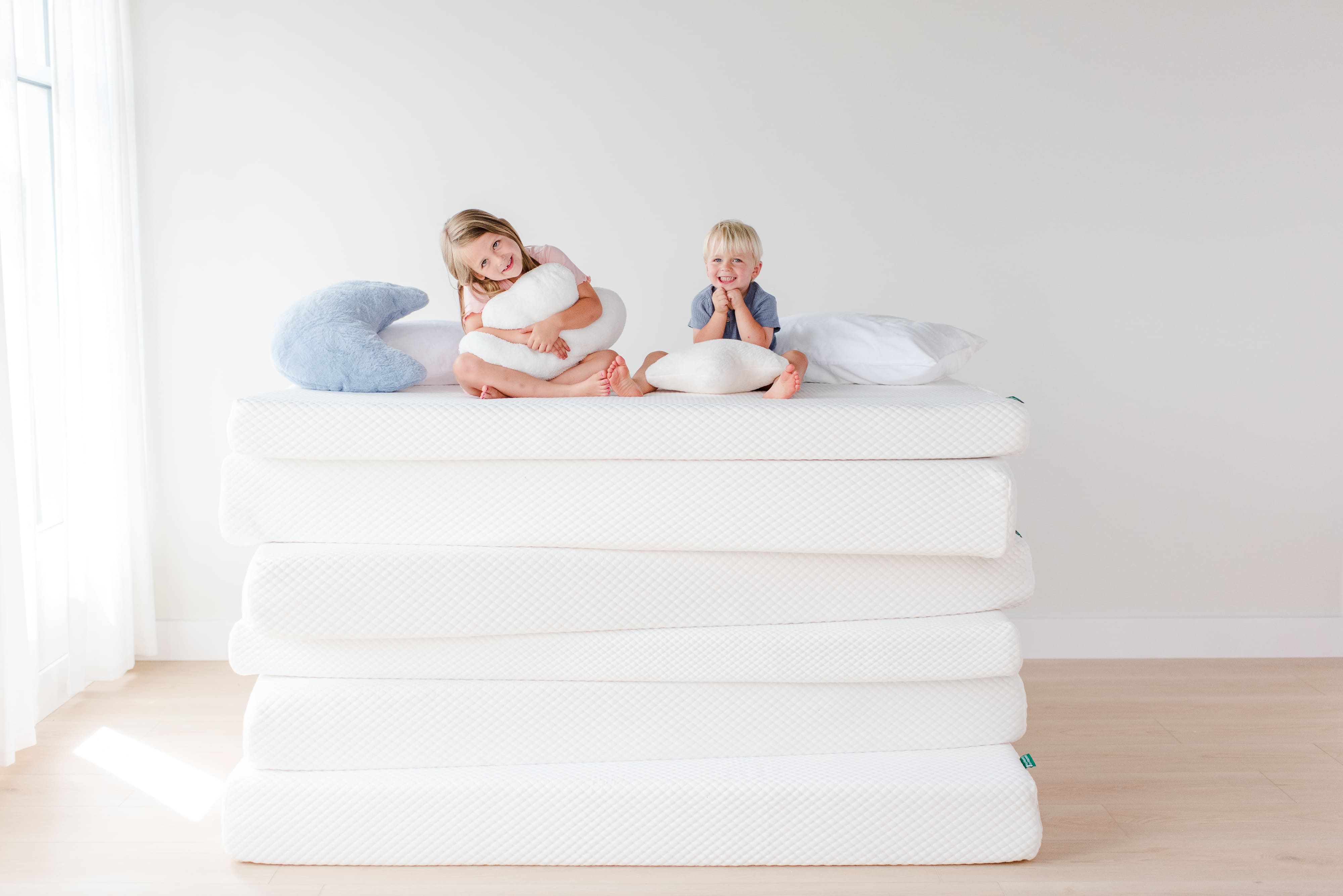A young girl and boy are sitting on a stack of mattresses, surrounded by a variety of pillows.