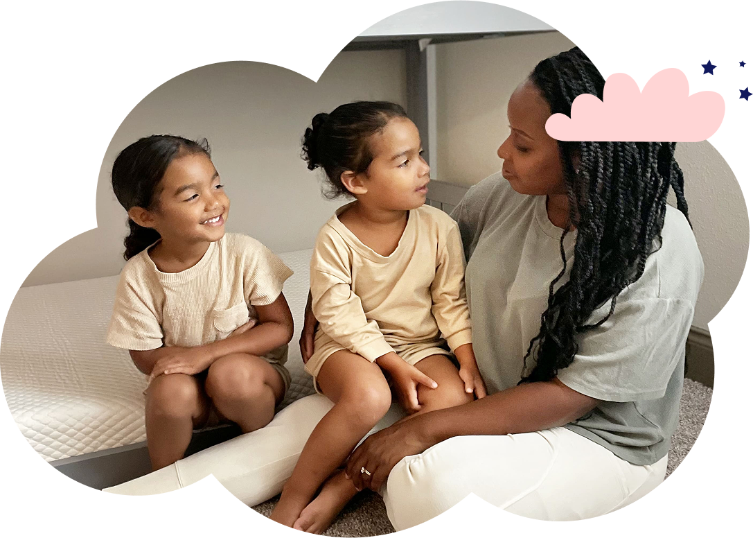 A mother sitting with her two daughters on the floor next to their bed. The mother has one daughter in her lap, the other next to her on the bottom bunk bed, smiling.