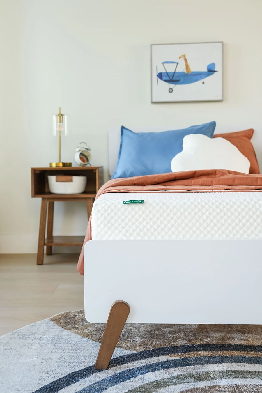 Bed with white quilted mattress, rust blanket, blue and cloud-shaped pillows, next to a wooden nightstand with a lamp and clock.