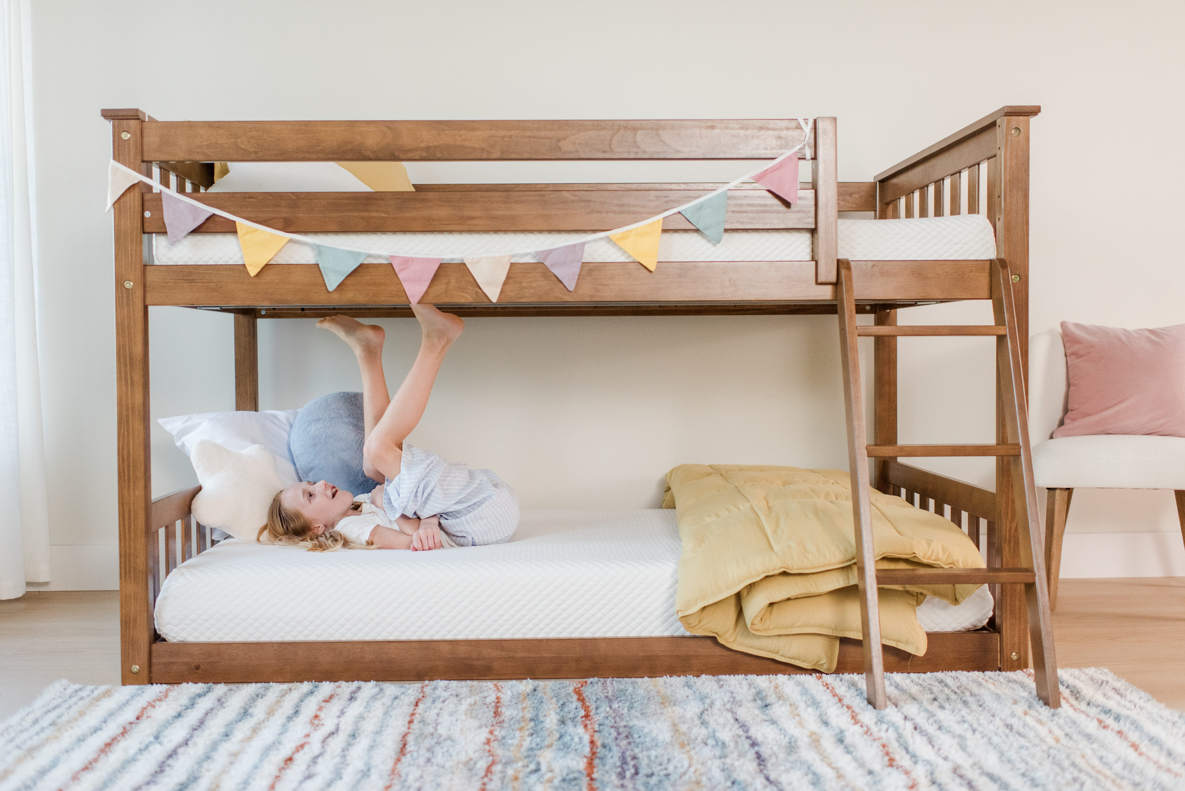 A young girl lying on the bottom bunk of a brown bunk bed decorated with colorful flags, kicking her feet in the air and smiling.
