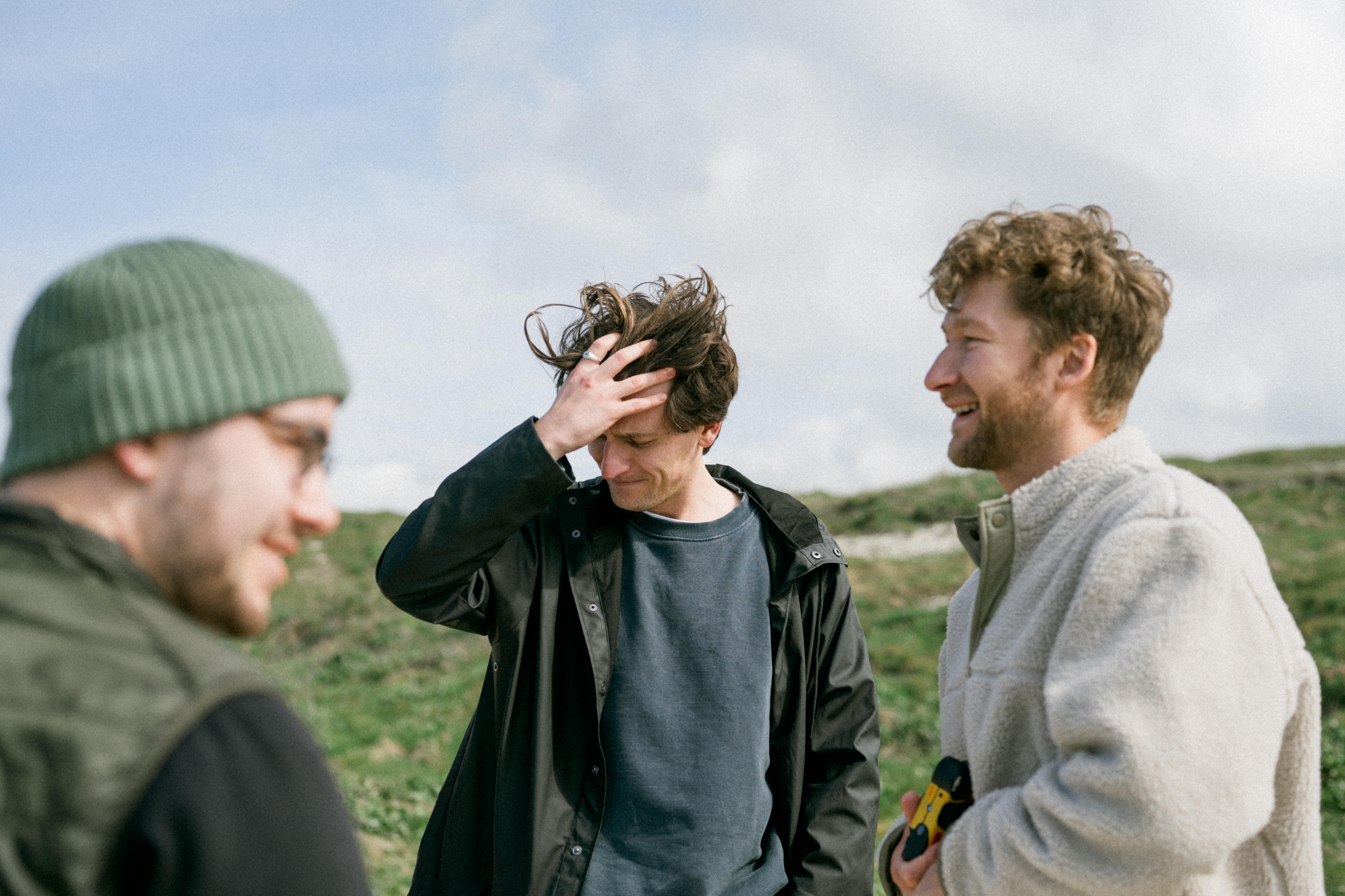 Three men outdoors, one adjusting hair, another smiling, with grassy background.