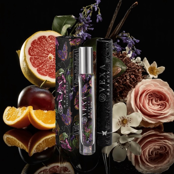 Perfume bottle with floral packaging surrounded by fruits and flowers on a reflective surface.