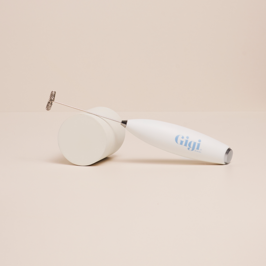A white Gigi handheld milk frother resting on a cylindrical block against a plain, light-colored background.