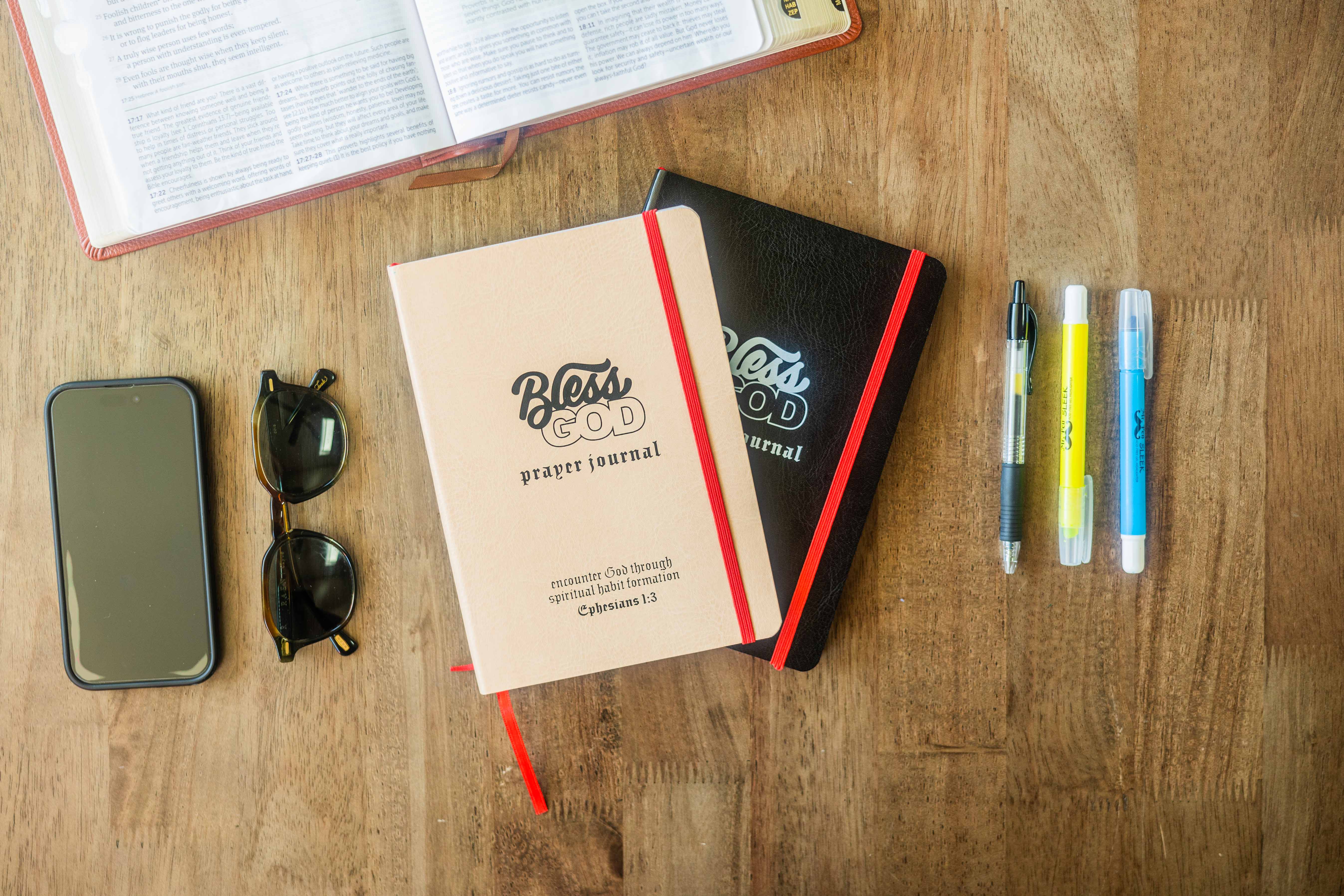 Open Bible, phone, sunglasses, journals, pens, and highlighters on a wooden table.