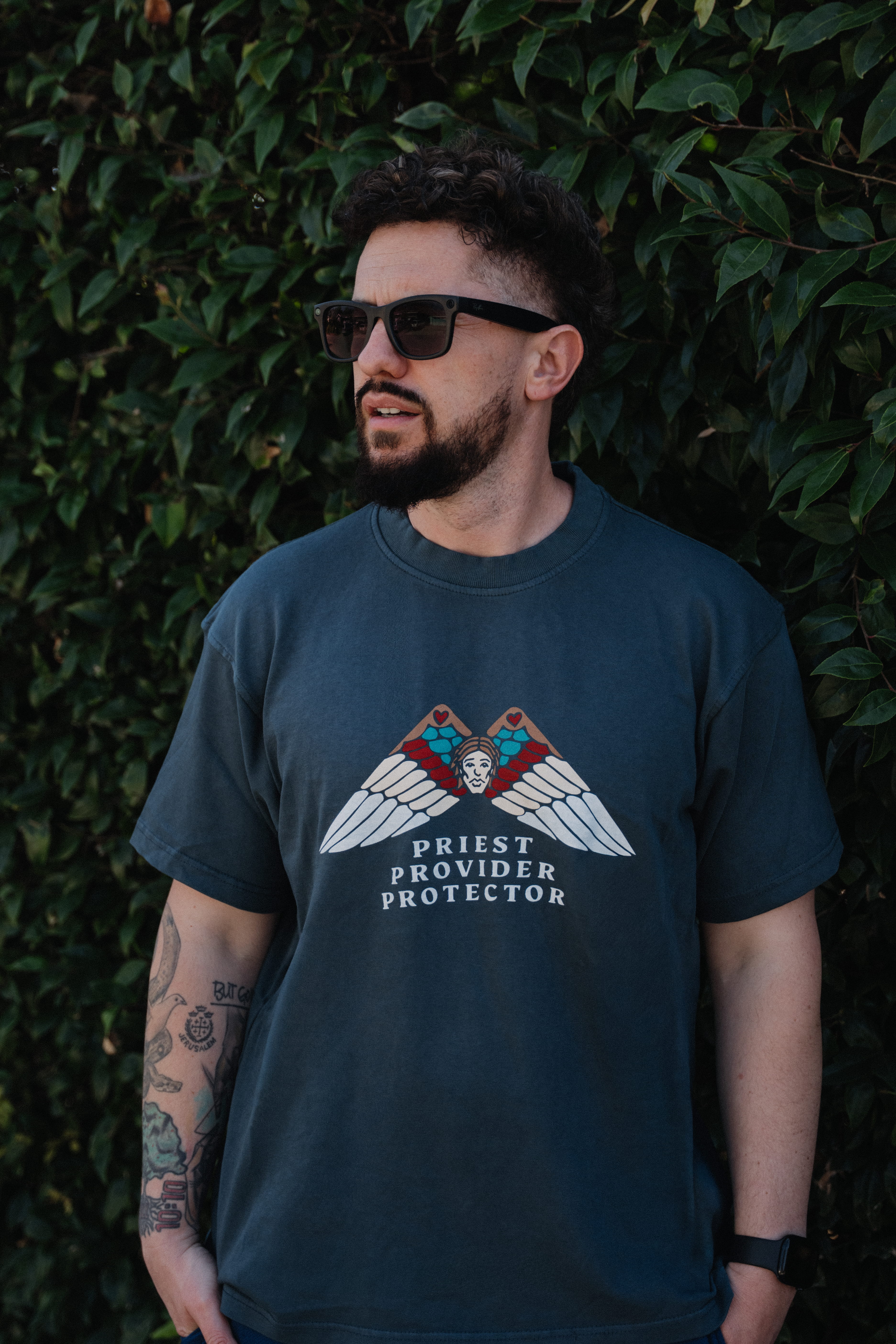 A man in sunglasses and a graphic tee that reads 'Priest Provider Protector' stands before a hedge.