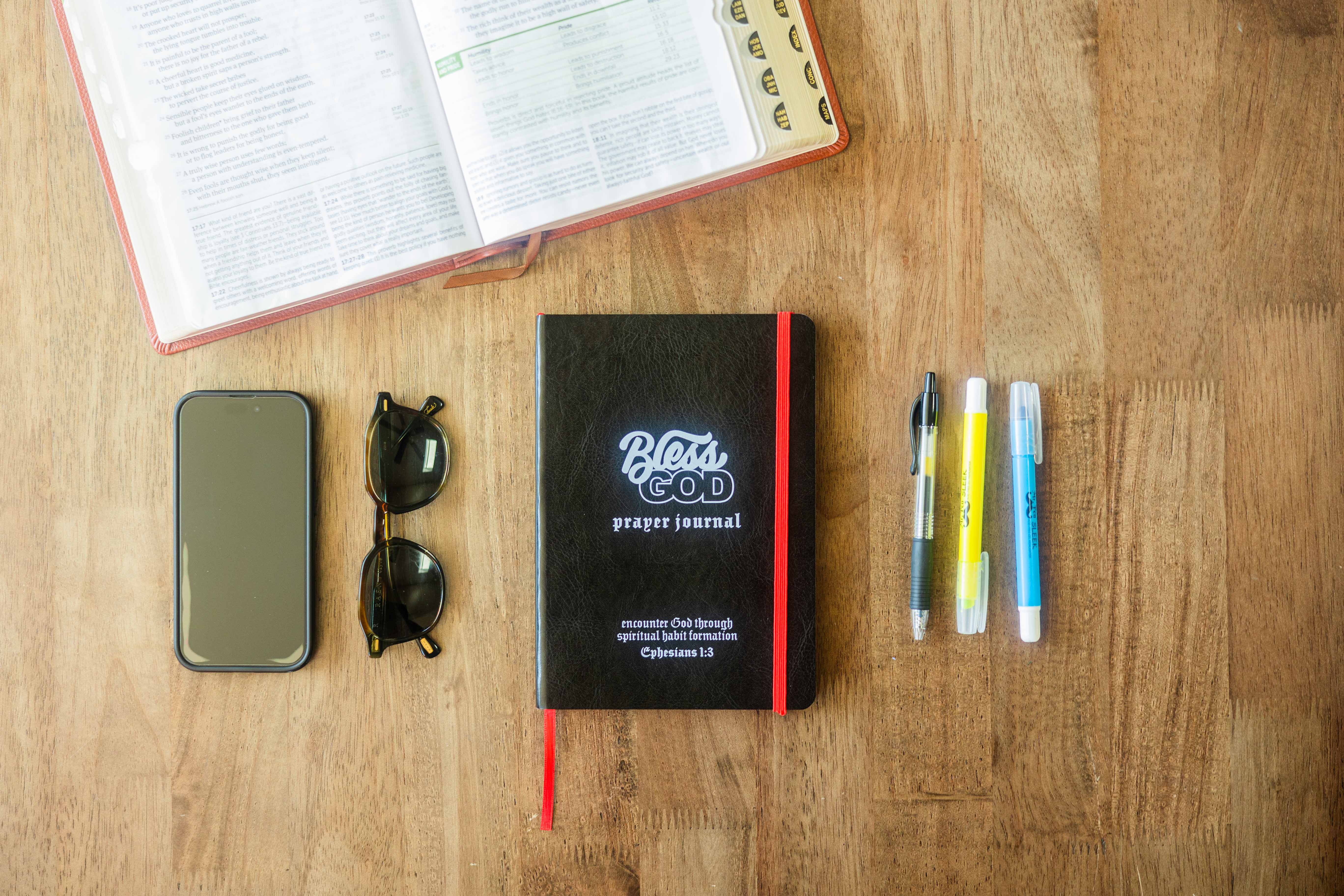 A Bible, prayer journal, phone, sunglasses, and pens on a wooden table.