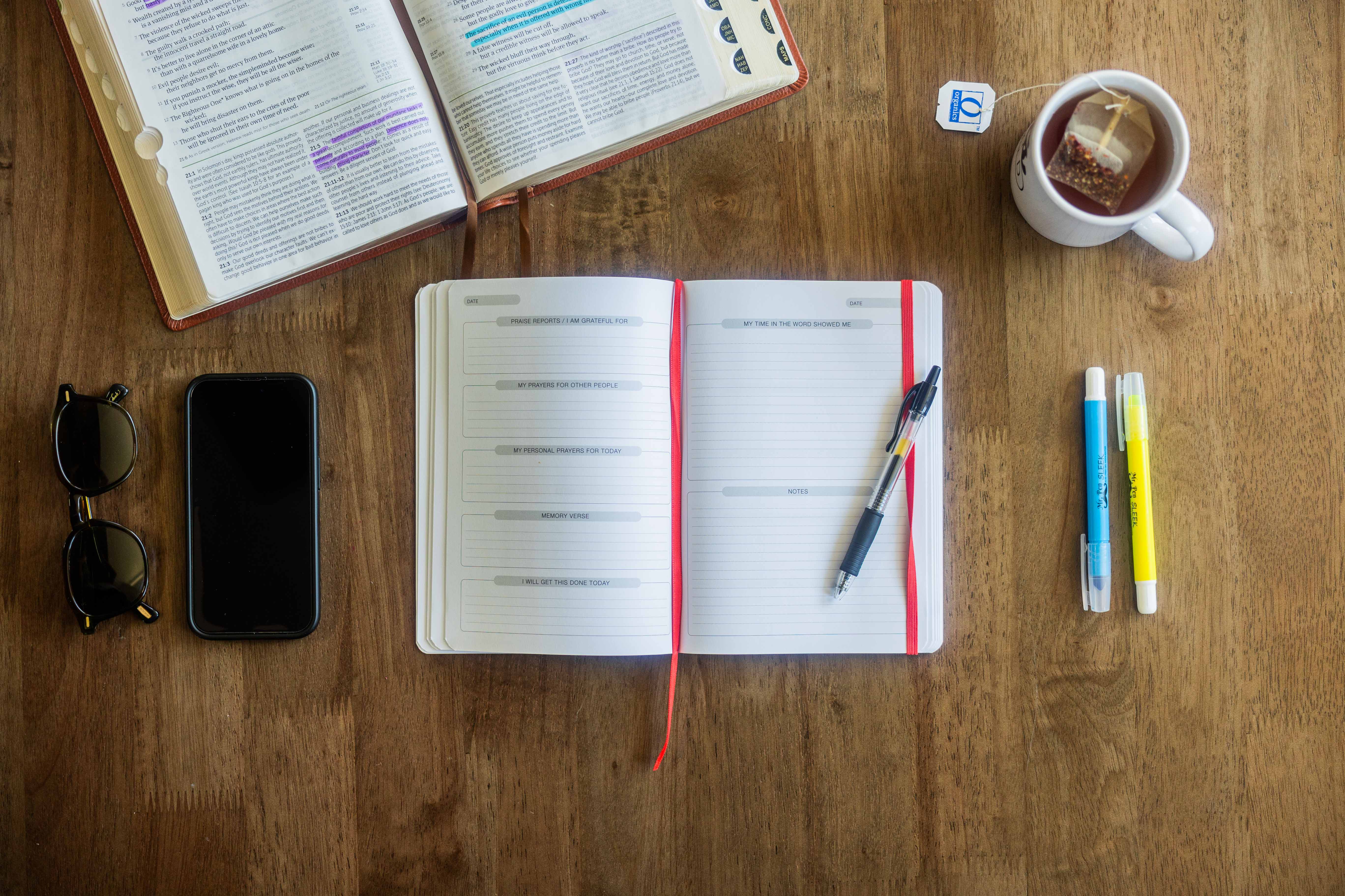 Open book, journal, pen, phone, sunglasses, tea on a wooden table.