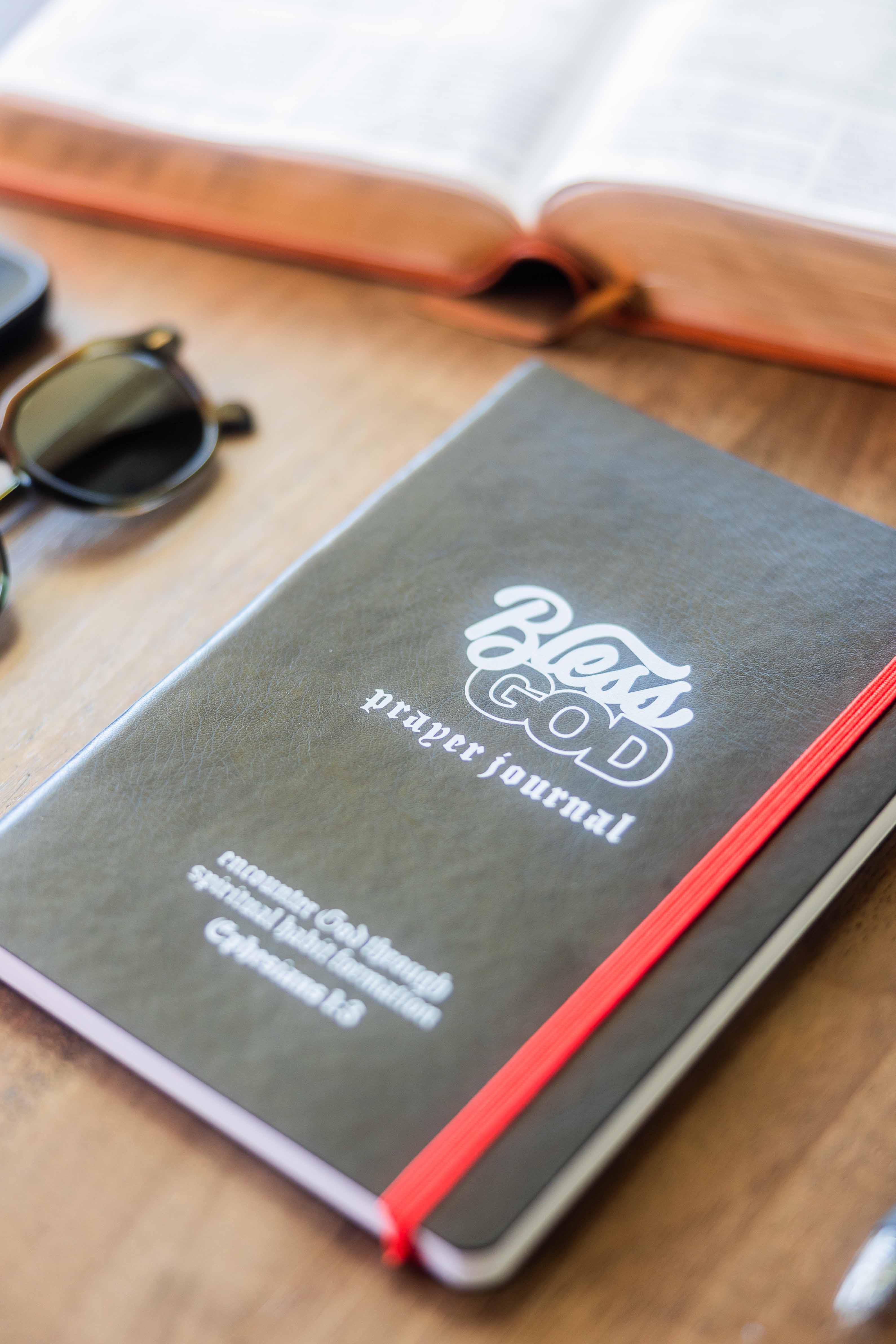 Prayer journal with red elastic band, open book, and sunglasses on a table.