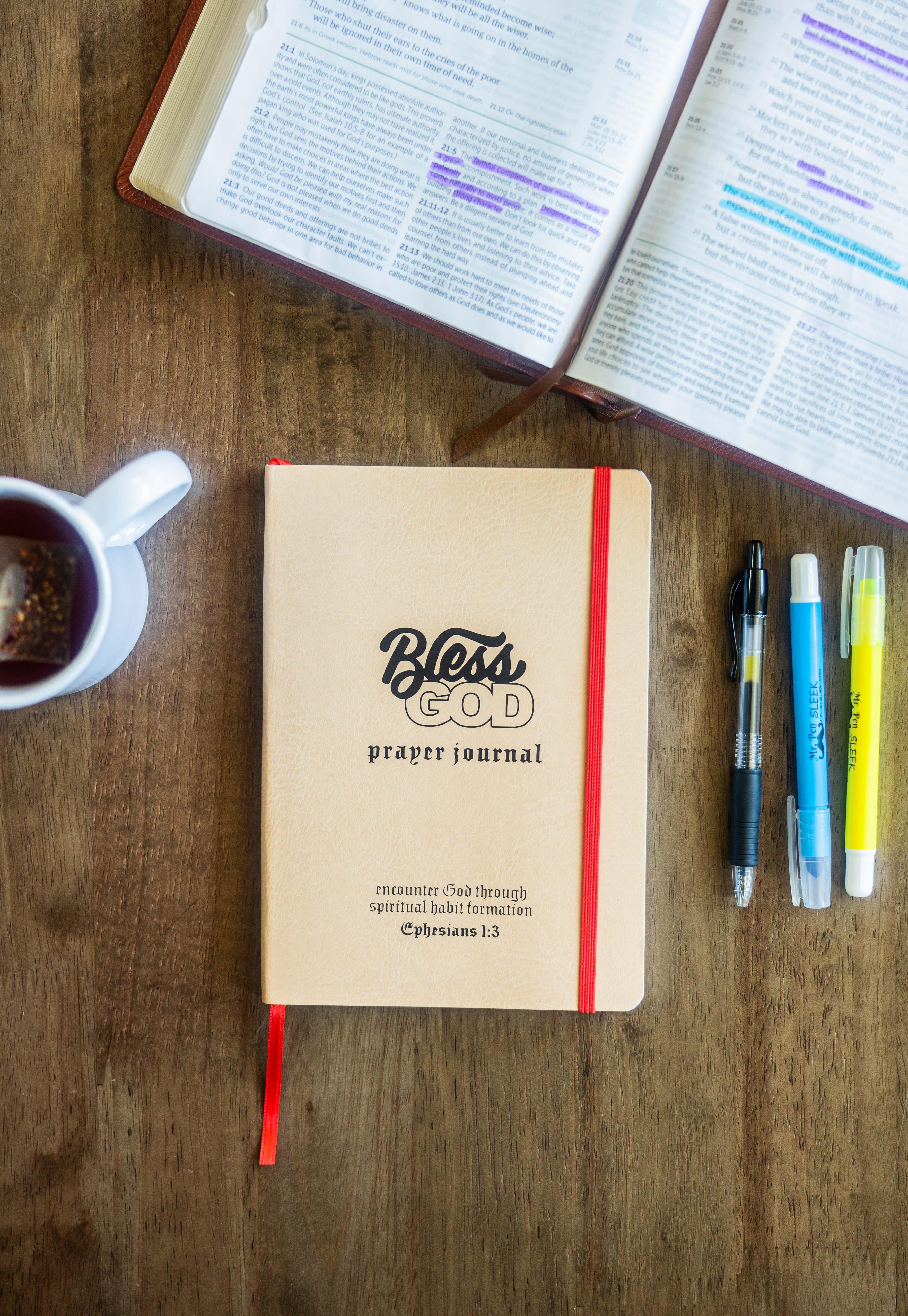 Prayer journal, pens, highlighters, open book, and tea on a wooden table.