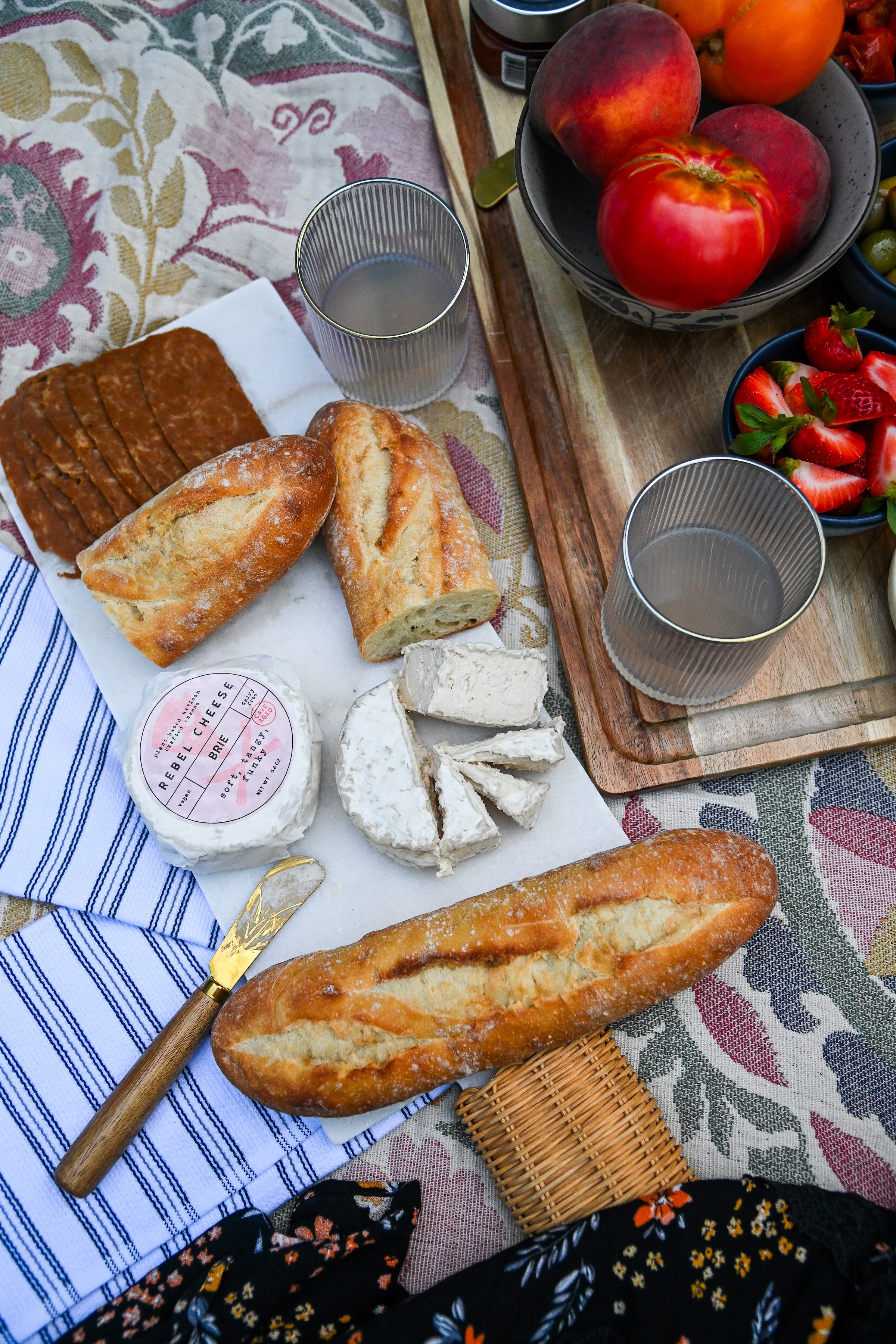 Picnic setup with bread, cheese, fruits, and drinks on a patterned cloth.