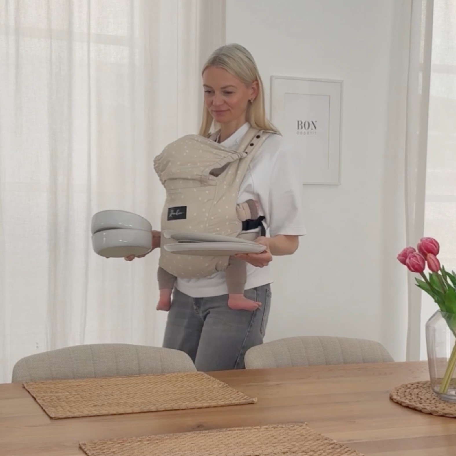 Woman carrying baby in a carrier, holding plates in a dining room.