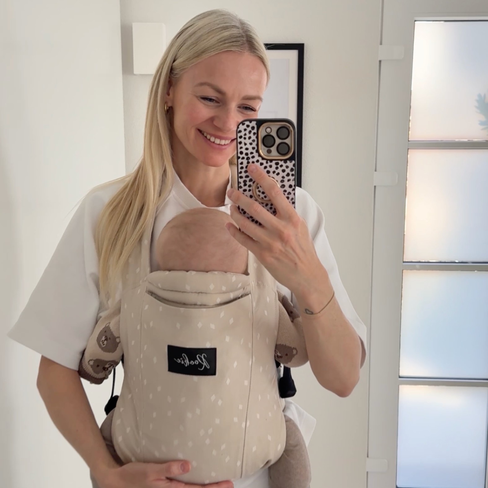Woman taking a mirror selfie with a baby in a beige carrier.