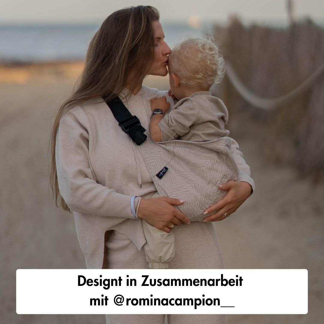 Person holding a child in a baby carrier, kissing their head on a beach.