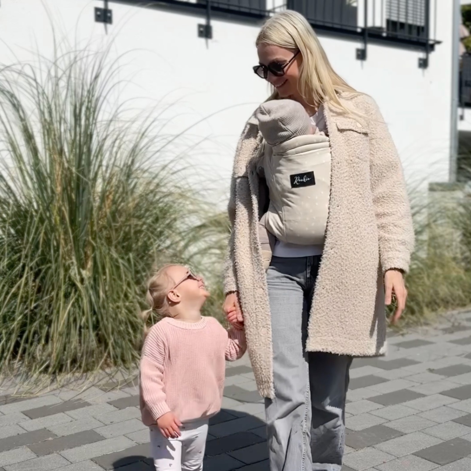 Woman with baby carrier and child walking outdoors, holding hands.