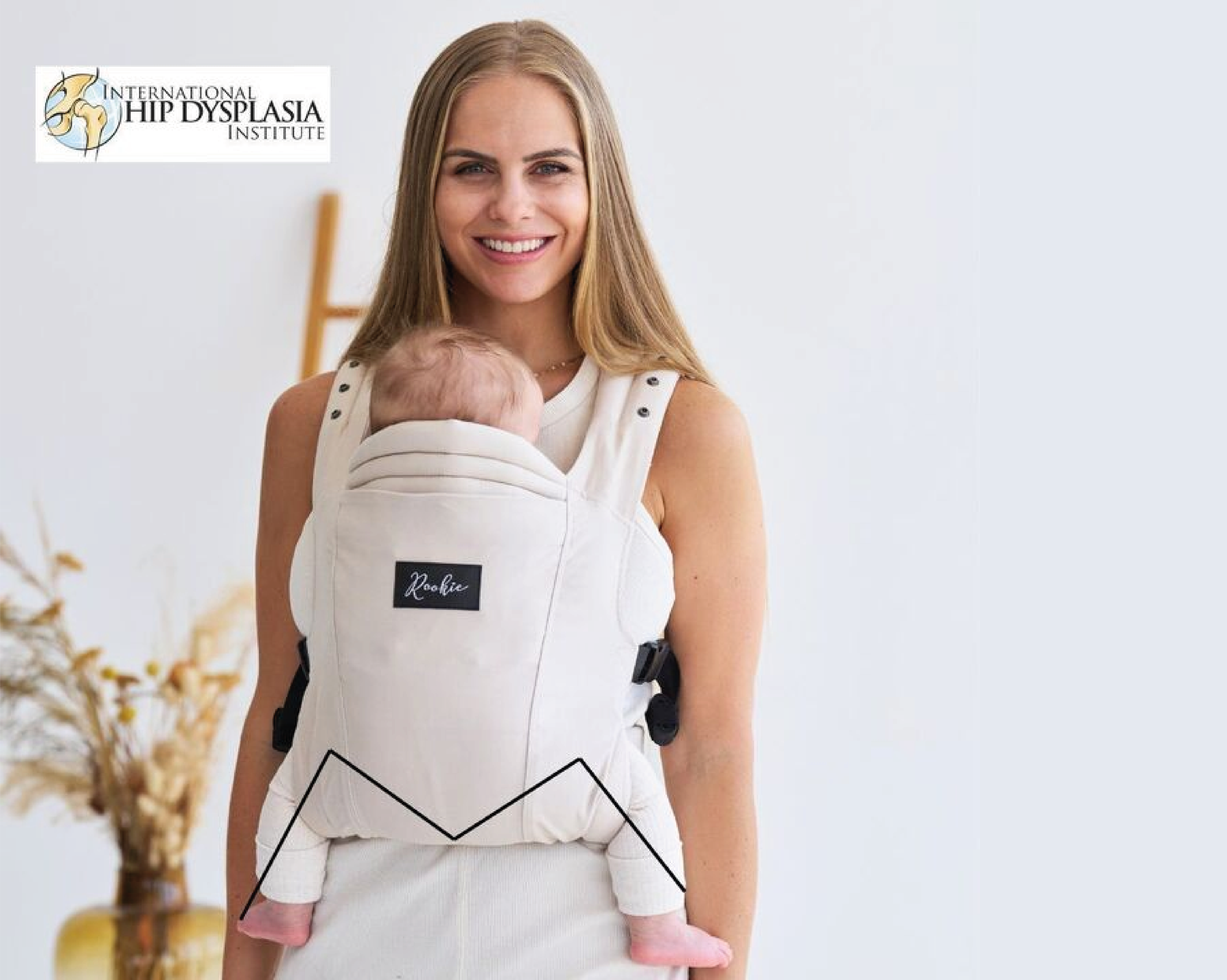Woman smiling, carrying a baby in a white carrier with a logo, indoors.