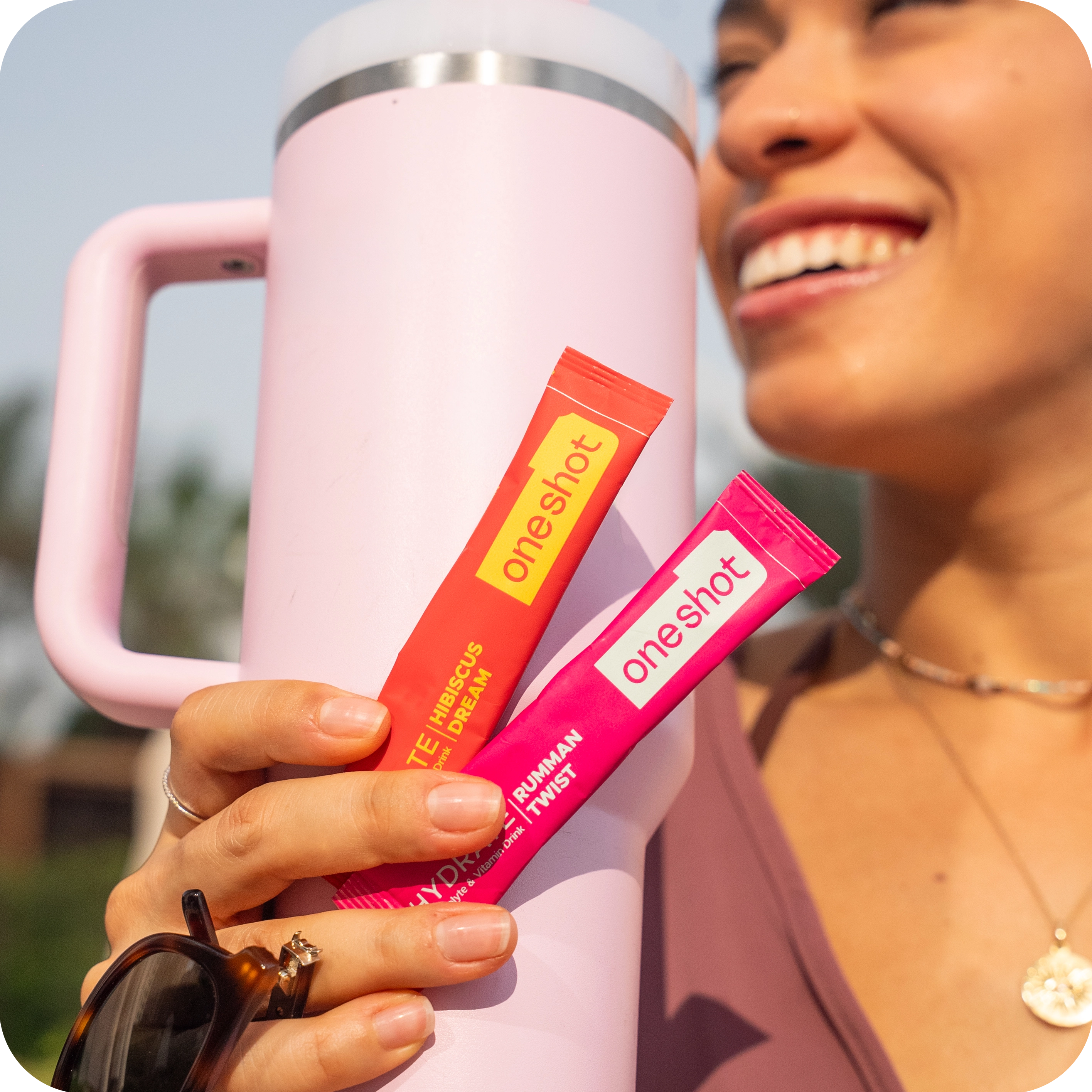 A smiling woman holds a pink water bottle and two 'one shot' drink mix packets outdoors.